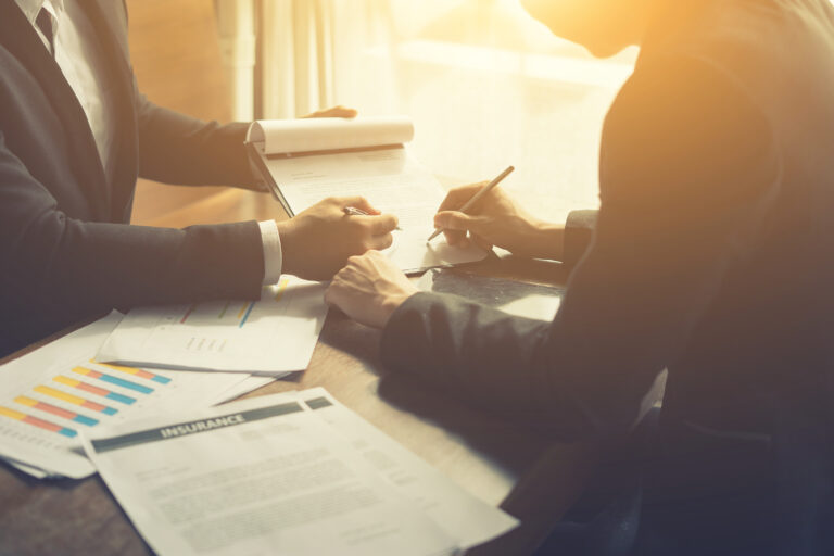 Two people in business attire signing paperwork, with an insurance document and a bar chart printout on the table