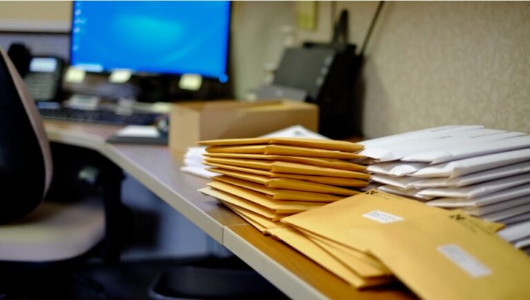 Stacks of manila and white envelopes on an HR desk beside a computer, ready for mailing ahead of a compliance deadline